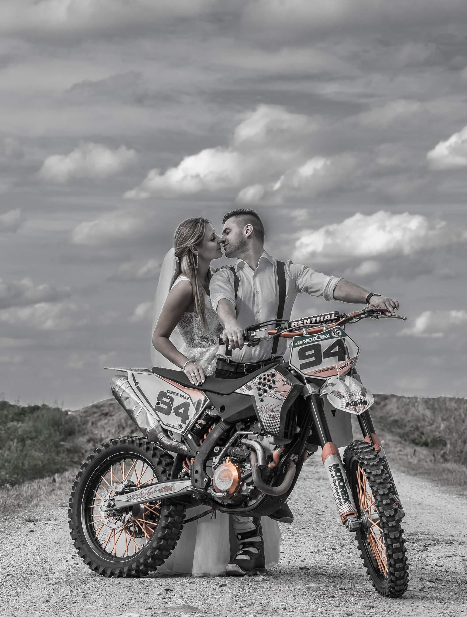 Para młoda w strojach ślubnych całuje się siedząc na motocyklu crossowym z numerem 94 na tle pochmurnego nieba. Zdjęcie w odcieniach szarości z pomarańczowymi akcentami.