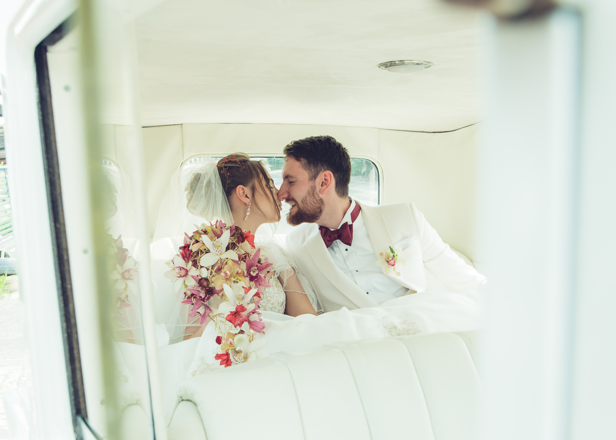 Panna młoda z bukietem z białych i różowych orchidei oraz pan młody w białym smokingu z bordową muchą, siedzący w białej limuzynie, zbliżają się do pocałunku.