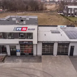 Widok z góry na budynek warsztatu samochodowego z dwoma częściami: stacja kontroli pojazdów RS Auto i myjnia ręczna z geometrią 3D wulkanizacją, panele słoneczne na dachu, szare otoczenie