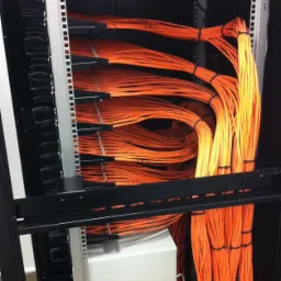 Pomarańczowe kable sieciowe uporządkowane w szafie rack, spięte czarnymi opaskami, widoczne urządzenia sieciowe w górnej części szafy.