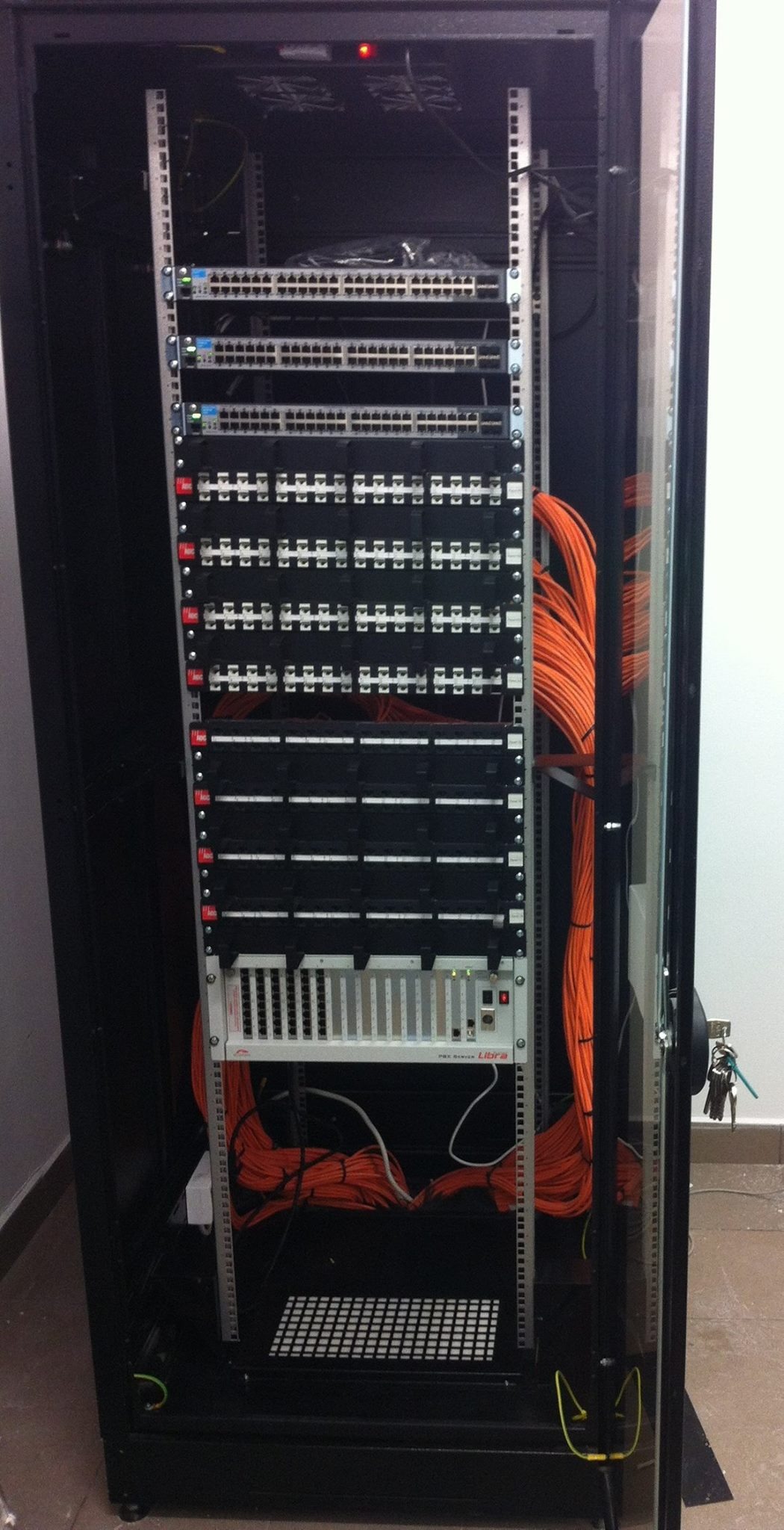 Szafa rack z patch panelem, switchami i okablowaniem w kolorze pomarańczowym, widoczne porty i diody sygnalizacyjne, klucze wiszące na drzwiach szafy.