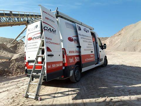 Samochód serwisowy z otwartymi drzwiami, z drabina opartą o tylne drzwi, na tle piaszczystego terenu, z widocznymi reklamami alarmów i monitoringu firmy INS-CAM z Kielc.