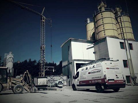 Biała furgonetka firmy INSCAM zaparkowana na placu budowy z betoniarnią w tle, obok koparka i wysoki dźwig.