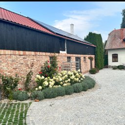 MKW-BUD - Dwukondygnacyjny budynek z cegły z czarną drewnianą górą i czerwoną dachówką, panele słoneczne na dachu, kwiaty i kostka brukowa na podwórku.