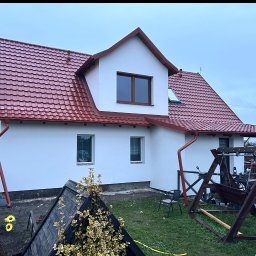 MKW-BUD - Biały dom z czerwoną dachówką i rynnami, widok z ogrodu. Nowe okna, świeżo pomalowana elewacja, zadbany trawnik. Huśtawka ogrodowa w tle.