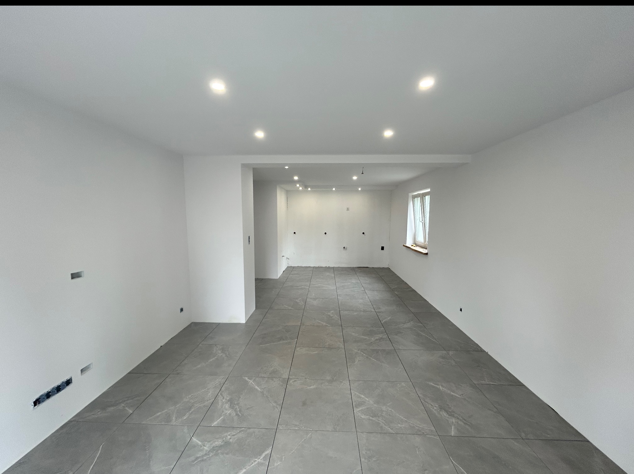 Wykończone wnętrze z szarymi płytkami podłogowymi imitującymi marmur. Białe ściany, sufit z oświetleniem punktowym. Widoczne puszki elektryczne w ścianach.