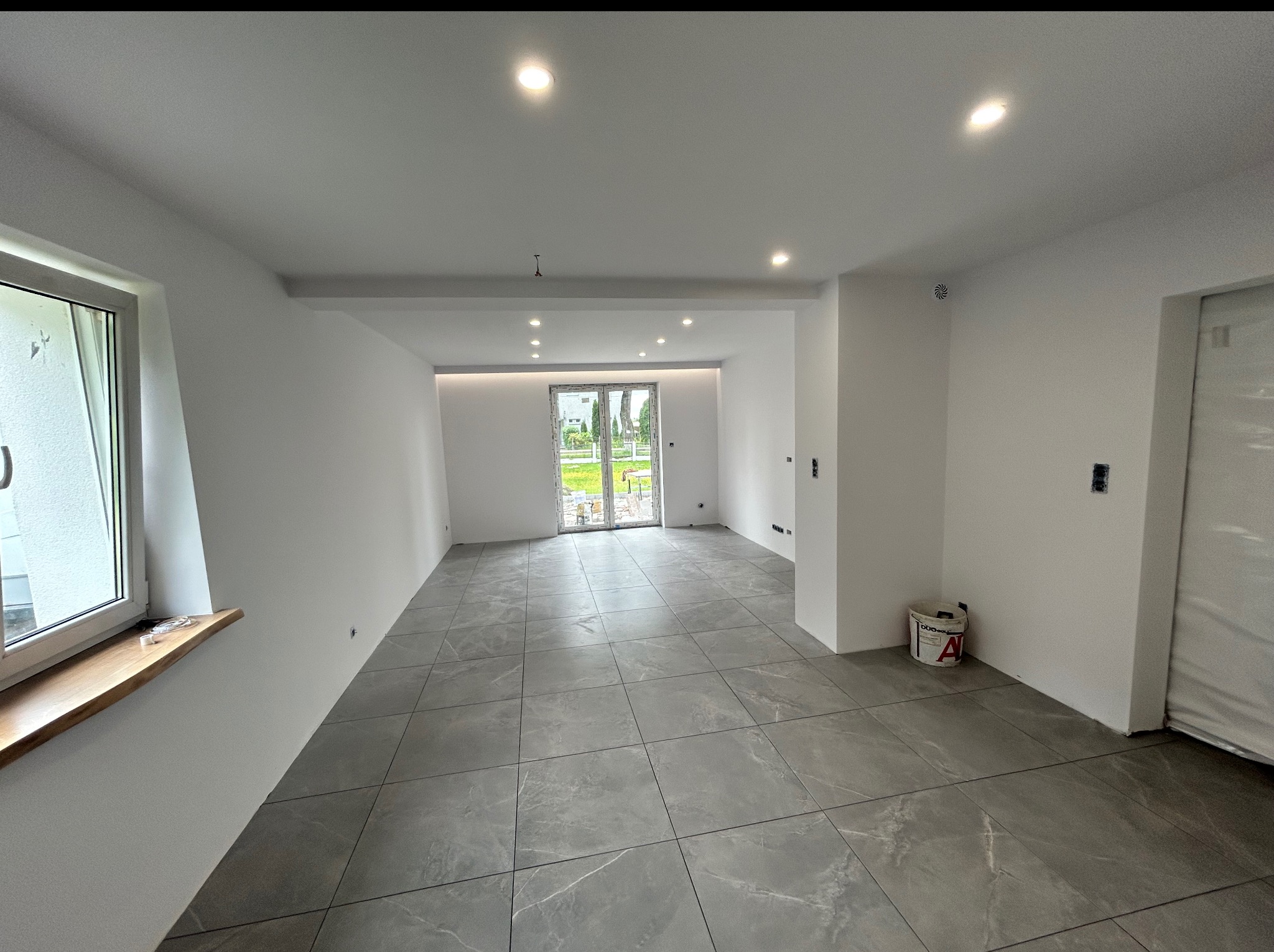 Jasne wnętrze po remoncie: szare płytki podłogowe z białymi żyłkami, białe ściany, okno z drewnianym parapetem, widok na ogród przez przeszklone drzwi.