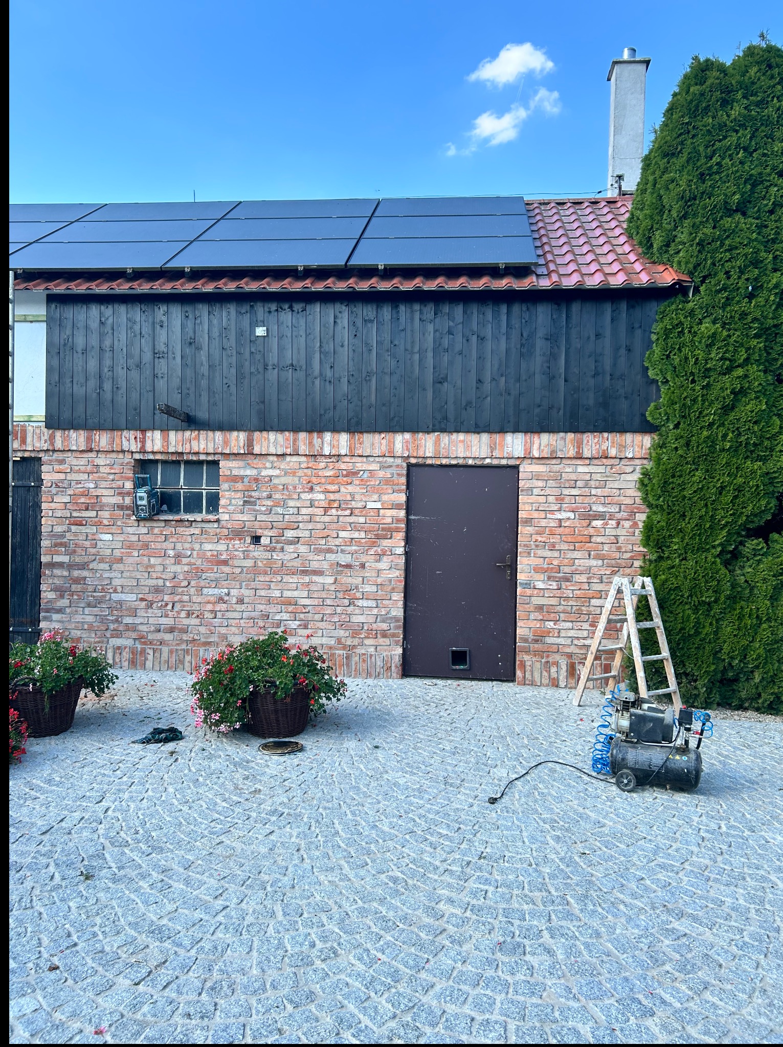 Budynek z cegły z panelami słonecznymi na dachu, drabina oparta o ścianę, kompresor na brukowanym podwórku. Widoczne kwiaty w donicach i ciemne, drewniane wykończenie.