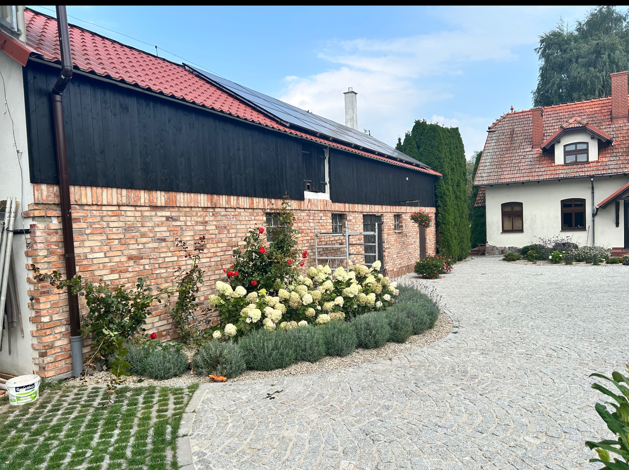 Dwukondygnacyjny budynek z cegły z czarną drewnianą górą i czerwoną dachówką, panele słoneczne na dachu, kwiaty i kostka brukowa na podwórku.