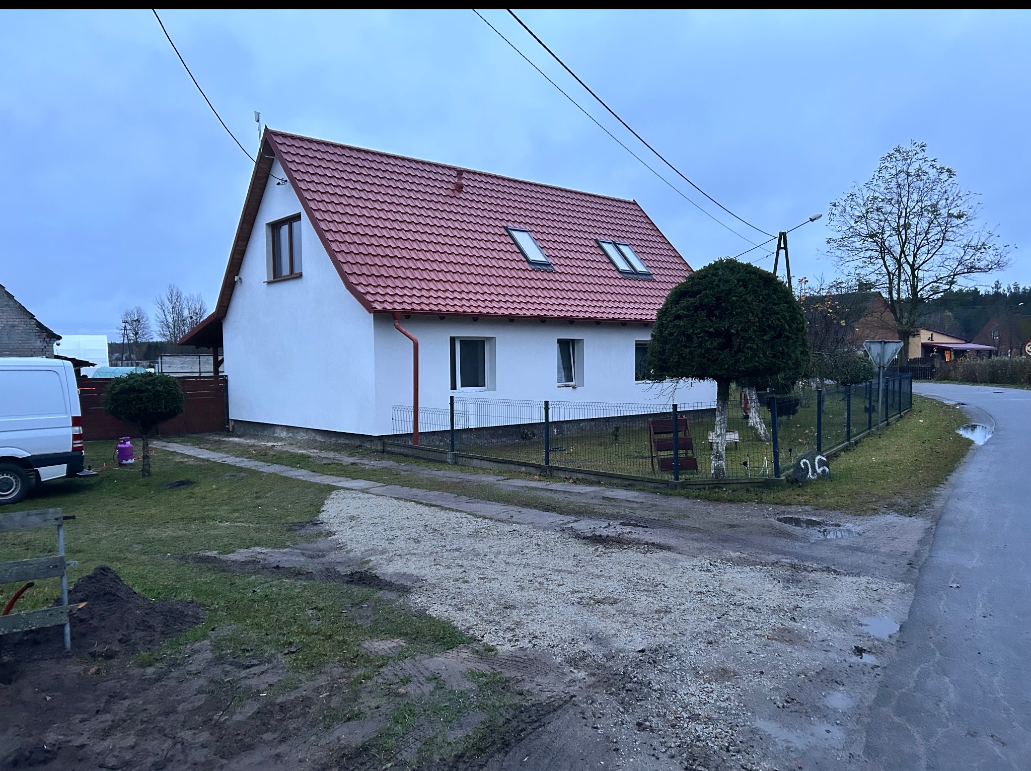 Biały dom z czerwoną dachówką i oknami dachowymi, otoczony zielenią i ogrodzeniem. Widok z zewnątrz, pochmurne niebo w tle. Świeżo pomalowana elewacja.