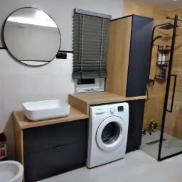 Nowoczesna łazienka z kabiną prysznicową typu walk-in, umywalką nablatową na szafce z szufladami, pralką i okrągłym lustrem na ścianie.