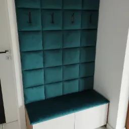 Tapicerowany panel ścienny w kolorze butelkowej zieleni z wieszakami i siedziskiem z szafką w przedpokoju, białe płytki podłogowe.