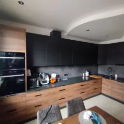 Nowoczesna kuchnia z drewnianymi frontami szafek dolnych i czarnymi szafkami górnymi, na blacie kuchennym stoją sprzęty AGD i owoce, widoczne dwa krzesła przy stole.