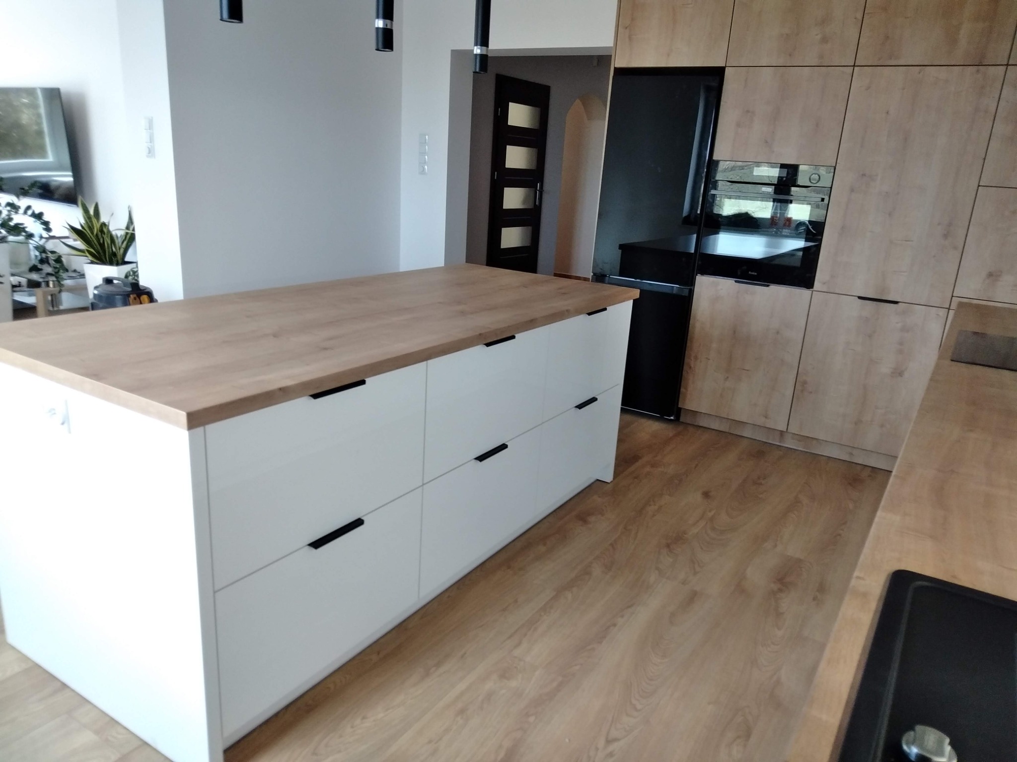 Wyspa kuchenna z białymi frontami i blatem imitującym jasne drewno, w tle zabudowa kuchenna w podobnej kolorystyce, czarny sprzęt AGD i drzwi z szybkami.