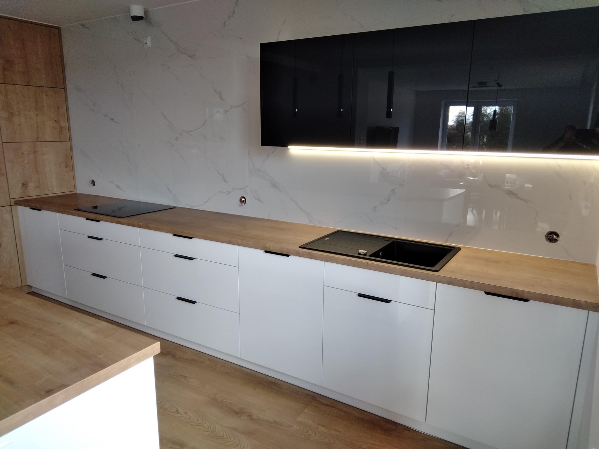 Nowoczesna kuchnia z białymi szafkami i drewnianym blatem, czarny zlewozmywak i płyta indukcyjna, marmurowe ściany, czarne szafki wiszące z oświetleniem LED.