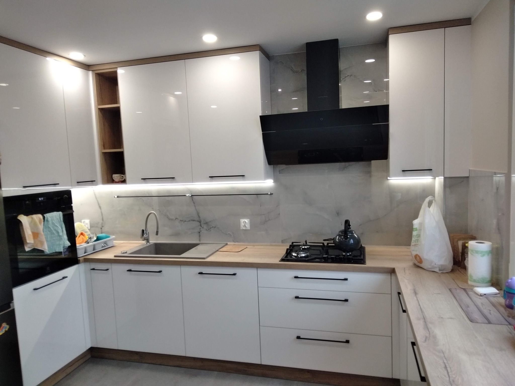 Nowoczesna kuchnia z białymi, lakierowanymi frontami, drewnianym blatem i czarnym okapem, oświetlona punktowo