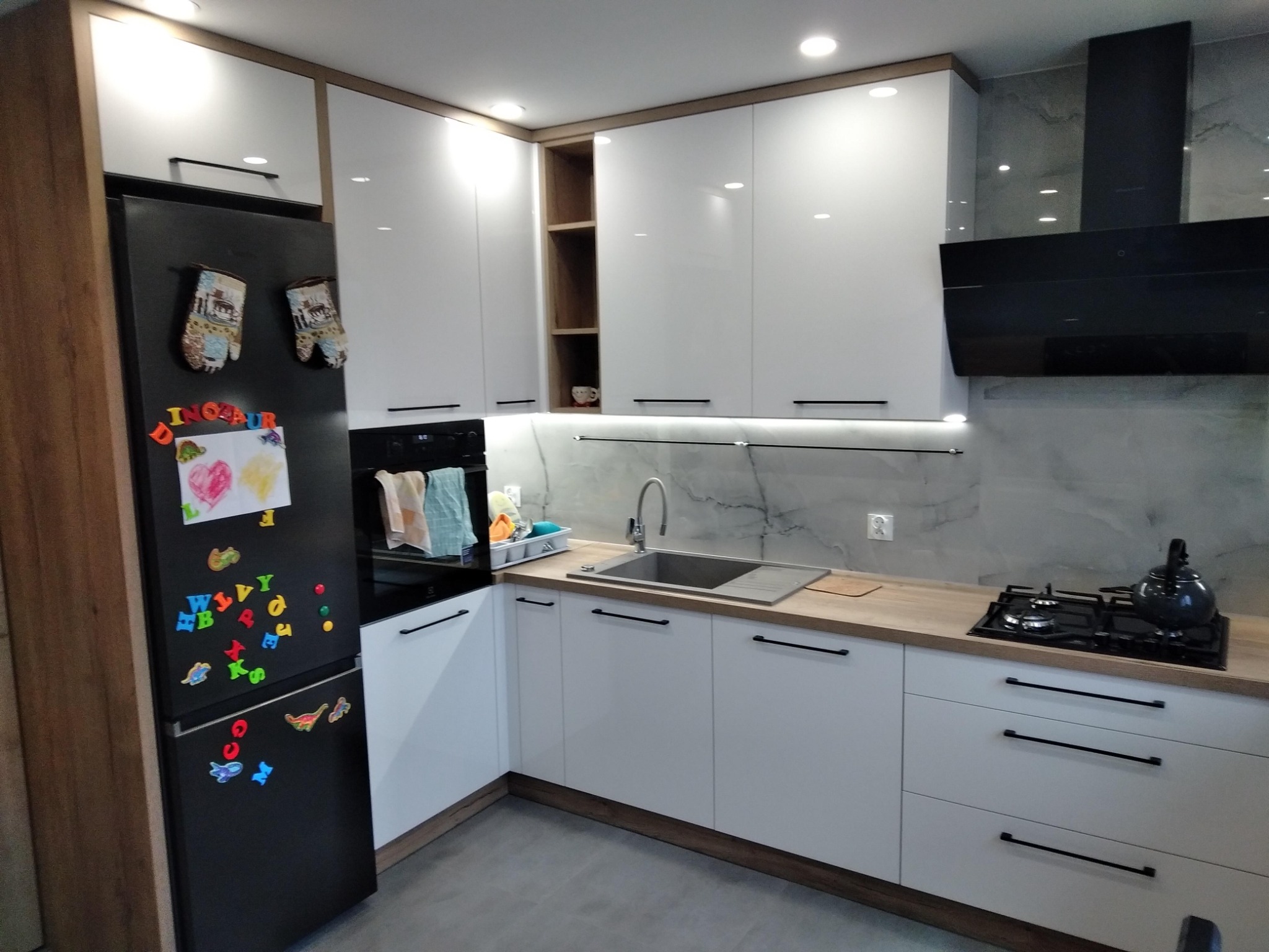 Biała kuchnia z drewnianymi akcentami, czarną lodówką ozdobioną magnesami i rysunkami dziecięcymi, oraz płytą gazową z czajnikiem.