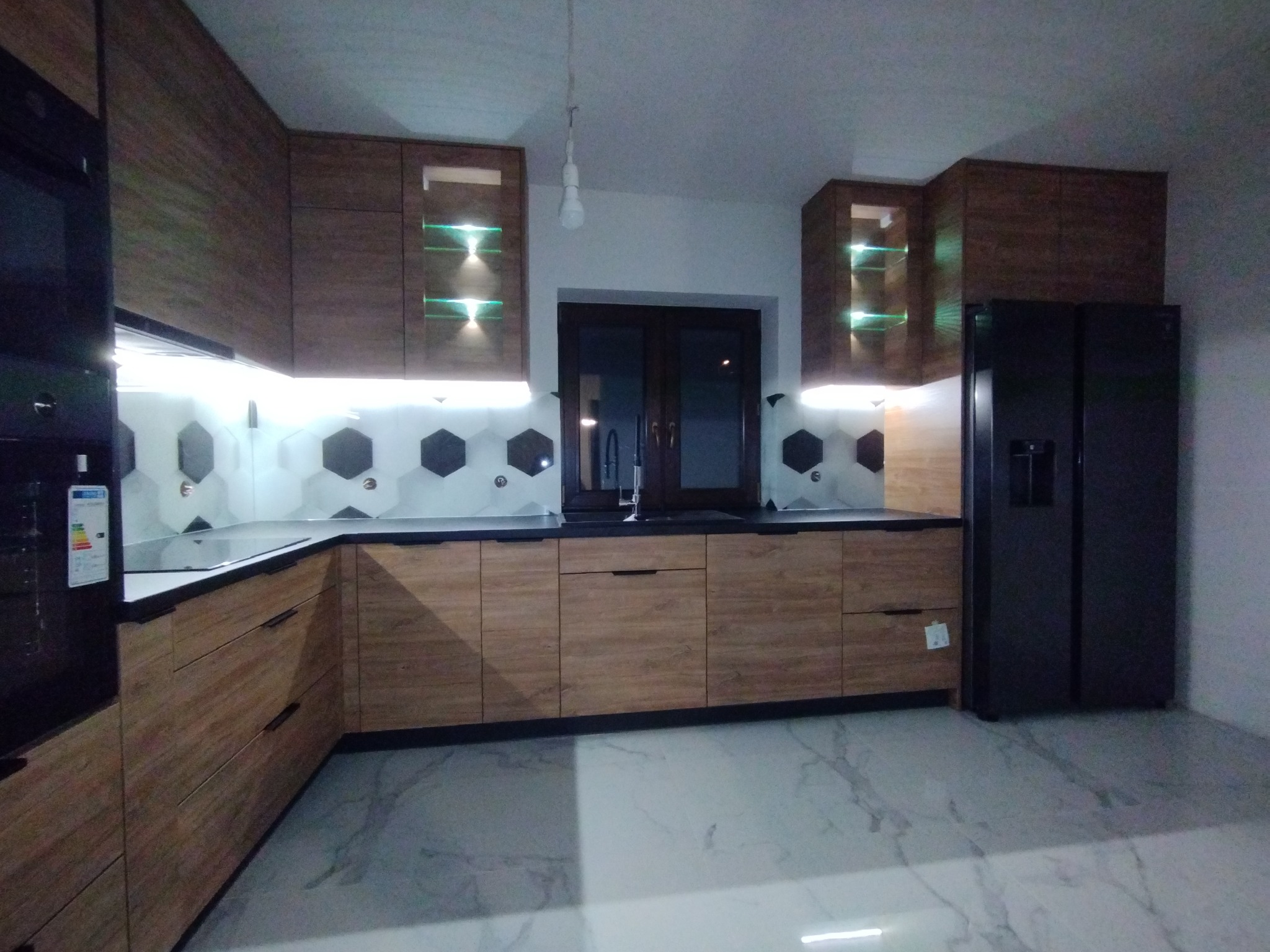 Nowoczesna kuchnia z drewnianymi szafkami, czarnym blatem, heksagonalnymi płytkami na ścianie i podświetlanymi półkami w górnych szafkach, uzupełniona czarną lodówką side-by-side oraz marmurową...