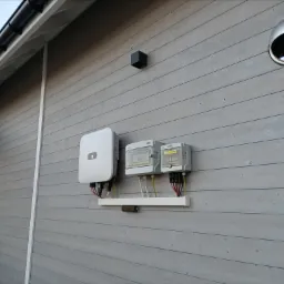 Biały inwerter i dwie szare skrzynki elektryczne zamontowane na jasnoszarej, drewnianej ścianie budynku, z widocznymi kablami i przewodami.