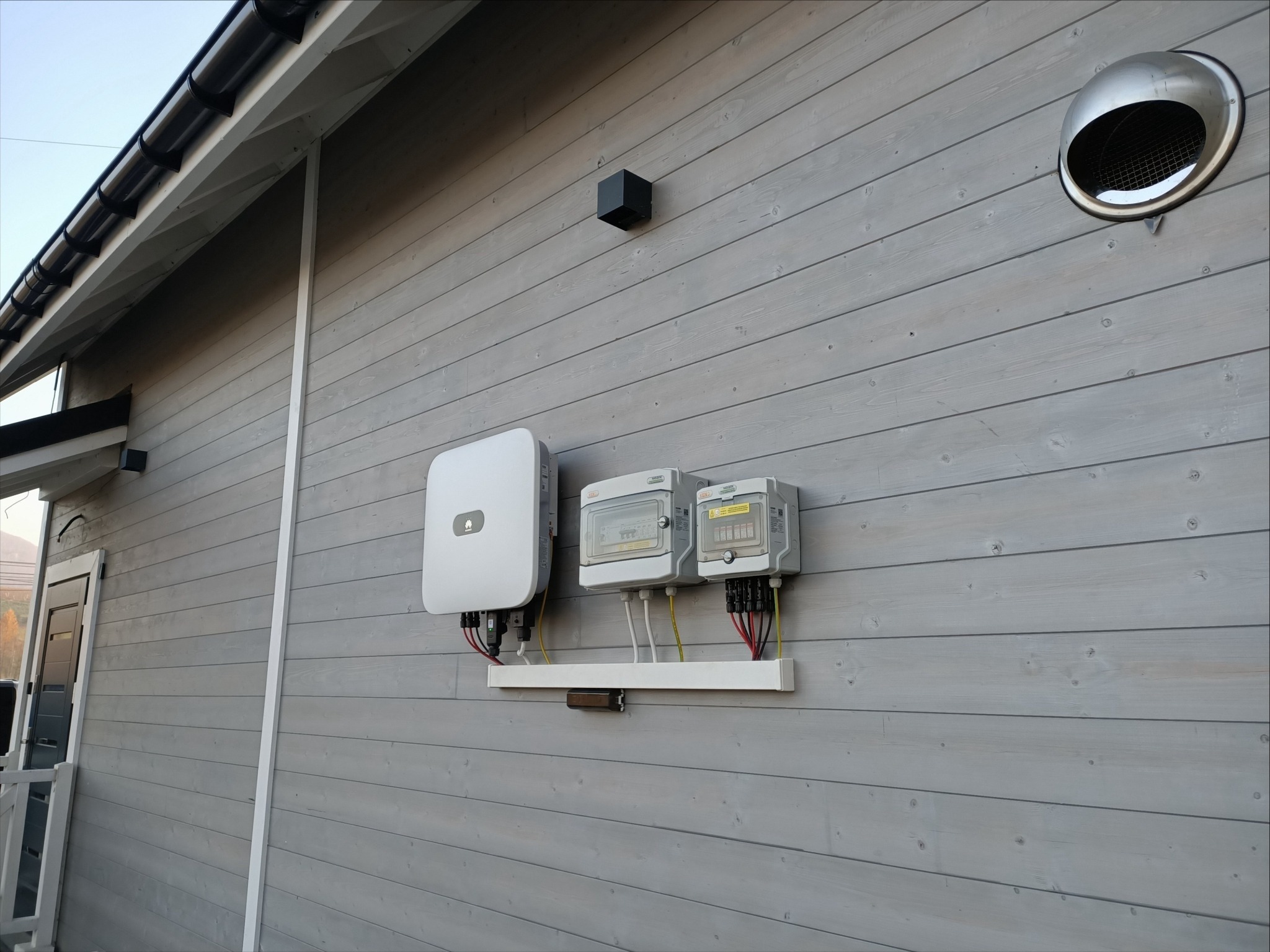Biały inwerter i dwie szare skrzynki elektryczne zamontowane na jasnoszarej, drewnianej ścianie budynku, z widocznymi kablami i przewodami.