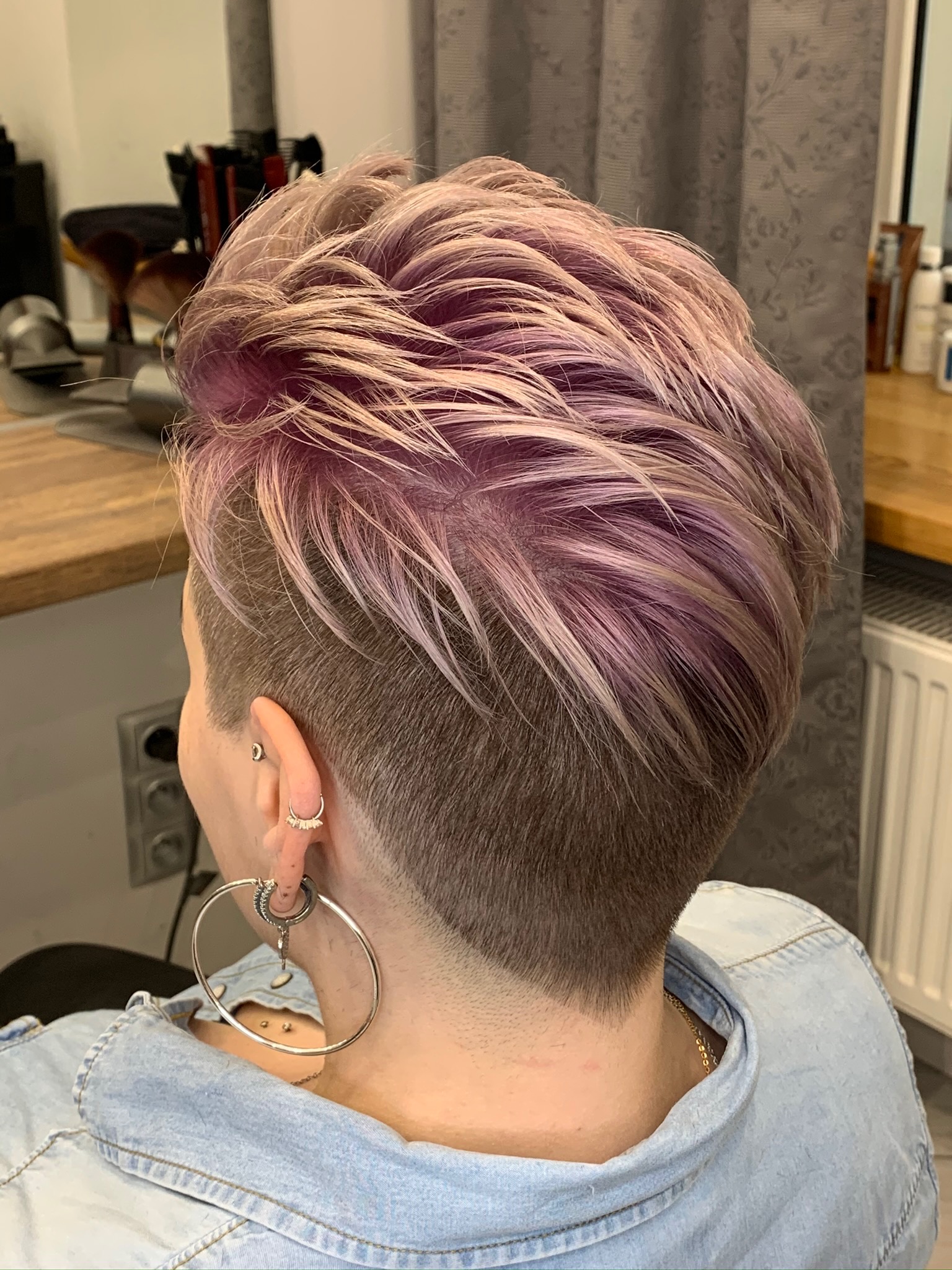 Krótkie, teksturowane włosy z różowo-blond koloryzacją, widoczne z tyłu głowy, z detalami kolczyków w uchu i jeansowej kurtki.