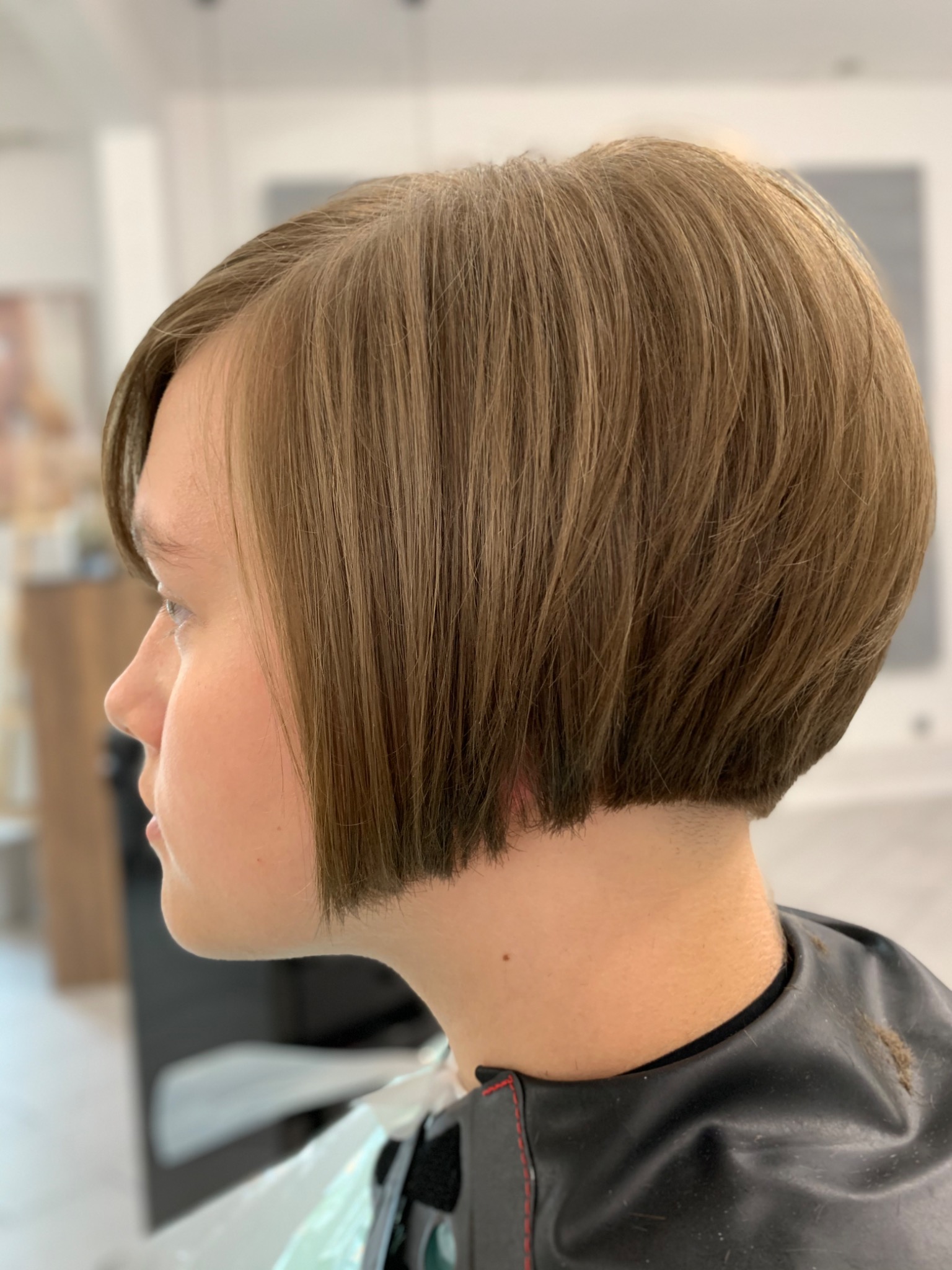 Profil kobiety z krótką, brązową fryzurą typu bob, widoczna peleryna ochronna, jasne tło.