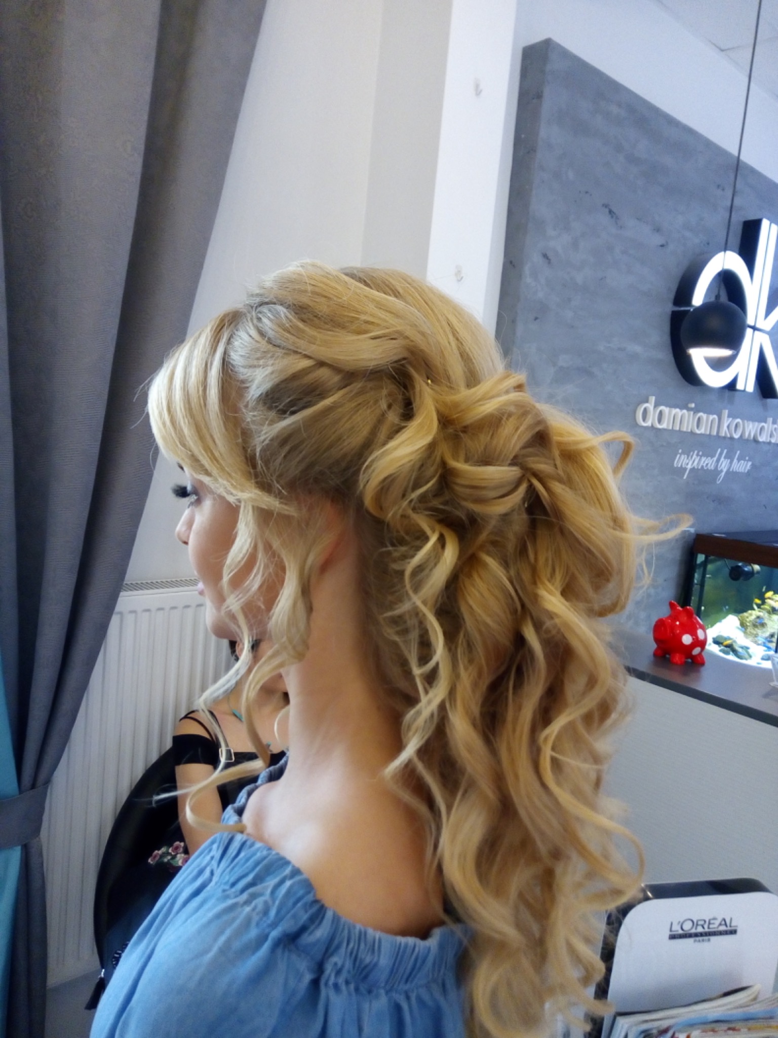 Upięcie ślubne z loków w odcieniach blondu, widok z tyłu głowy klientki w salonie, logo salonu na ścianie, akwarium w tle.