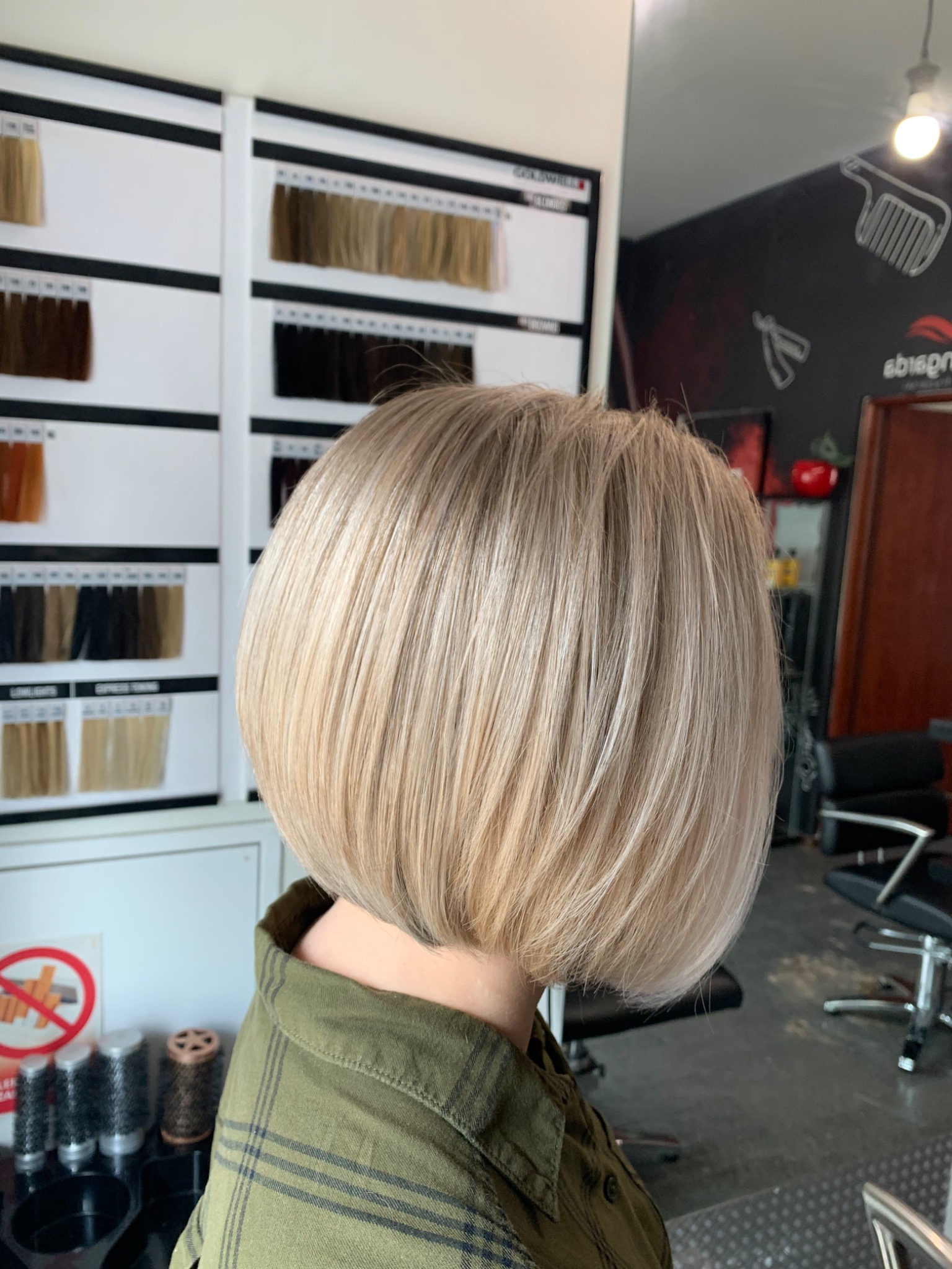 Krótkie, blond włosy po strzyżeniu bob, widok z tyłu, w tle paleta kolorów włosów i wnętrze salonu.