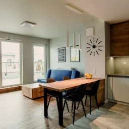 Jasne wnętrze mieszkania w Poznaniu z otwartą przestrzenią salonu z aneksem kuchennym, drewniany stół z czarnymi krzesłami, niebieska sofa, balkon z widokiem na osiedle.