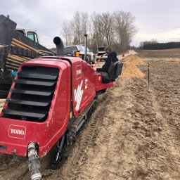 Fibertech sp zoo kom - Czerwona, gąsienicowa maszyna TORO DD2024 w trakcie pracy na polu, operator w słuchawkach, widoczna sterta piasku i rury na przyczepie, pochmurne niebo.