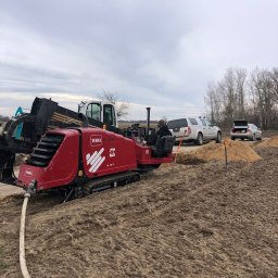 Fibertech sp zoo kom - Czerwona koparka Toro DD 2024 na gąsienicach, z wężem doprowadzającym wodę, w trakcie prac ziemnych na polu, z widocznym operatorem w okularach ochronnych, obok zaparkowane samochody.