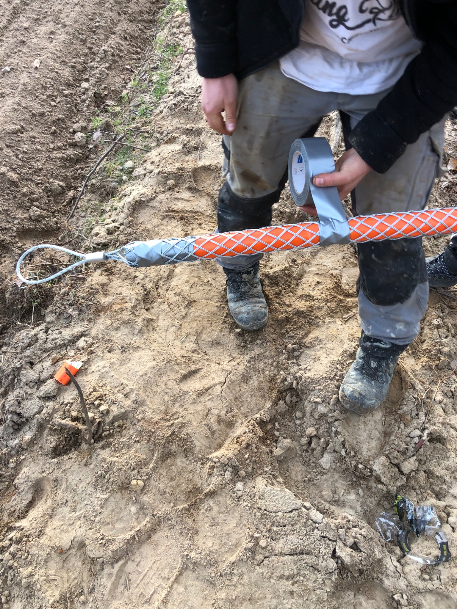 Mężczyzna zabezpiecza taśmą pomarańczowy kabel w siatce ochronnej na ziemi.