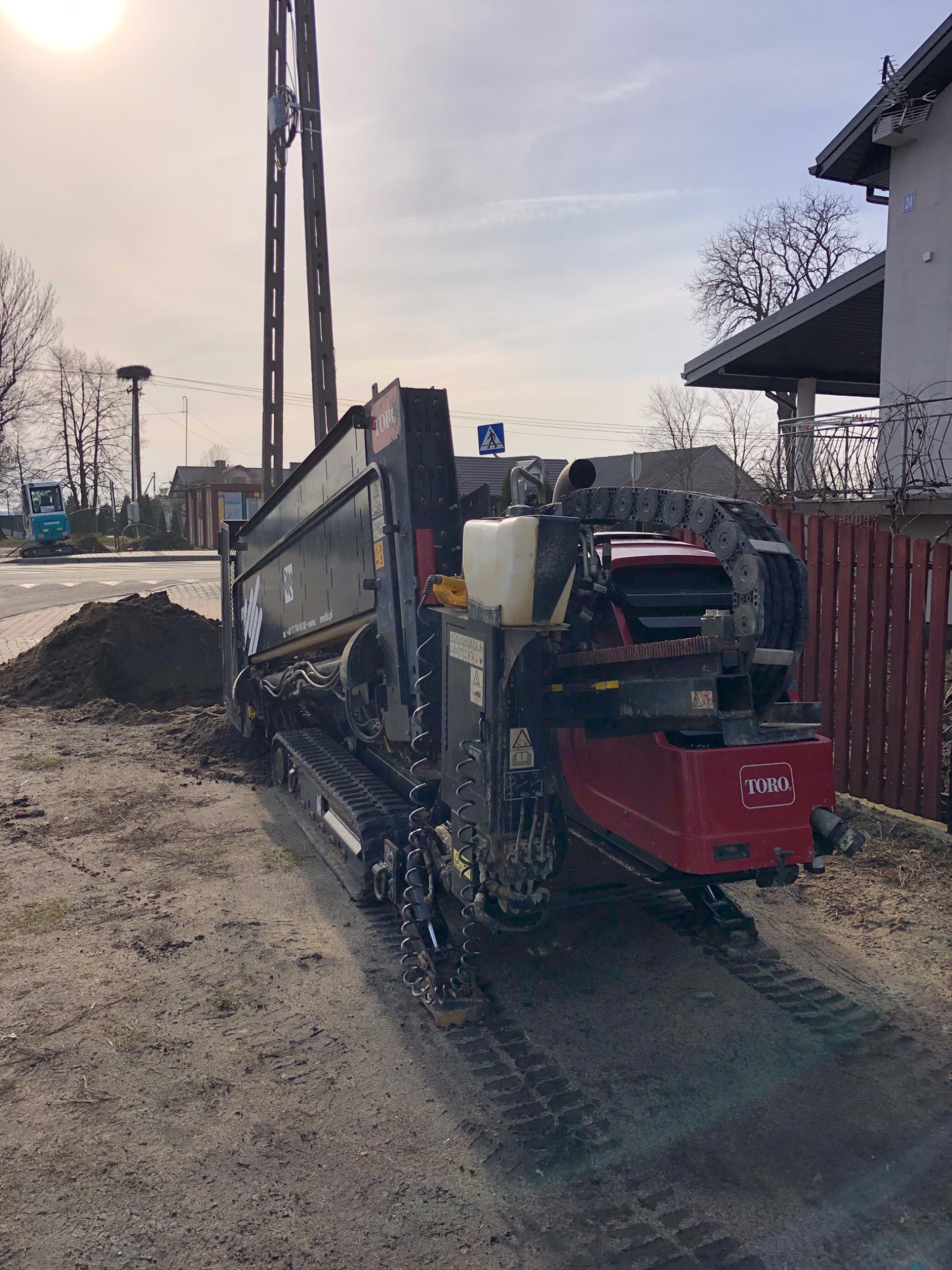 Gąsienicowa maszyna budowlana TORO na piaszczystym terenie, obok sterta ziemi i słup energetyczny, w tle budynki mieszkalne i częściowo widoczna koparka.