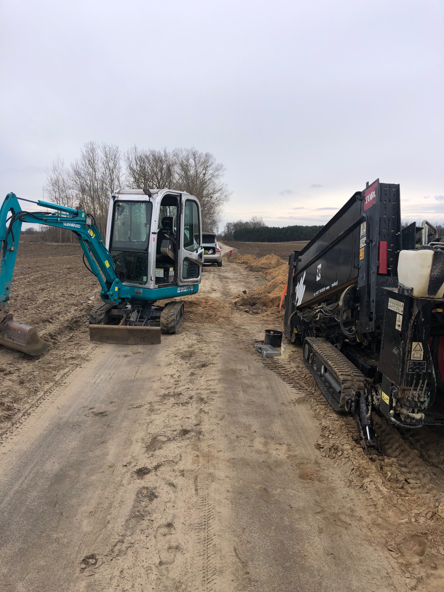 Turkusowa mini koparka Sunward i czarna maszyna Toro na gładkiej, piaszczystej drodze gruntowej, na tle pola i rzadkich drzew pod pochmurnym niebem.