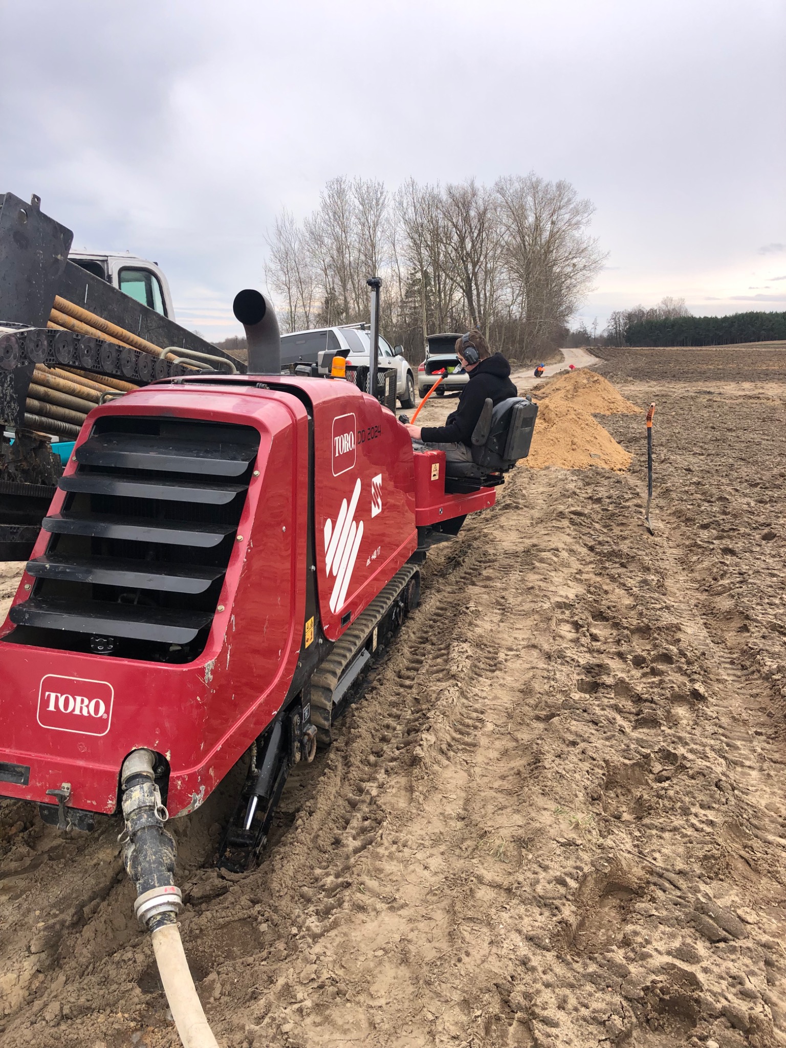 Czerwona, gąsienicowa maszyna TORO DD2024 w trakcie pracy na polu, operator w słuchawkach, widoczna sterta piasku i rury na przyczepie, pochmurne niebo.