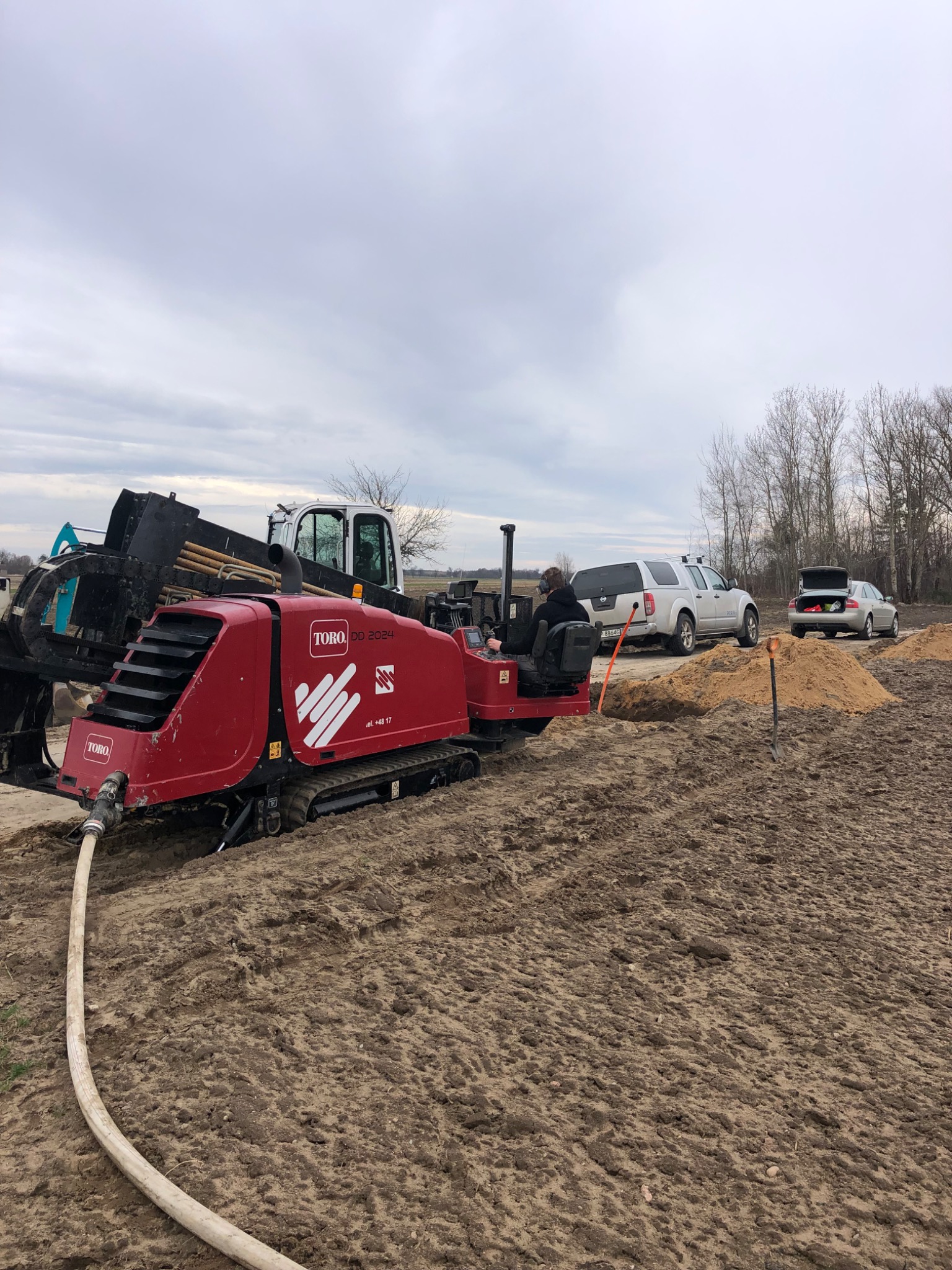 Czerwona koparka Toro DD 2024 na gąsienicach, z wężem doprowadzającym wodę, w trakcie prac ziemnych na polu, z widocznym operatorem w okularach ochronnych, obok zaparkowane samochody.