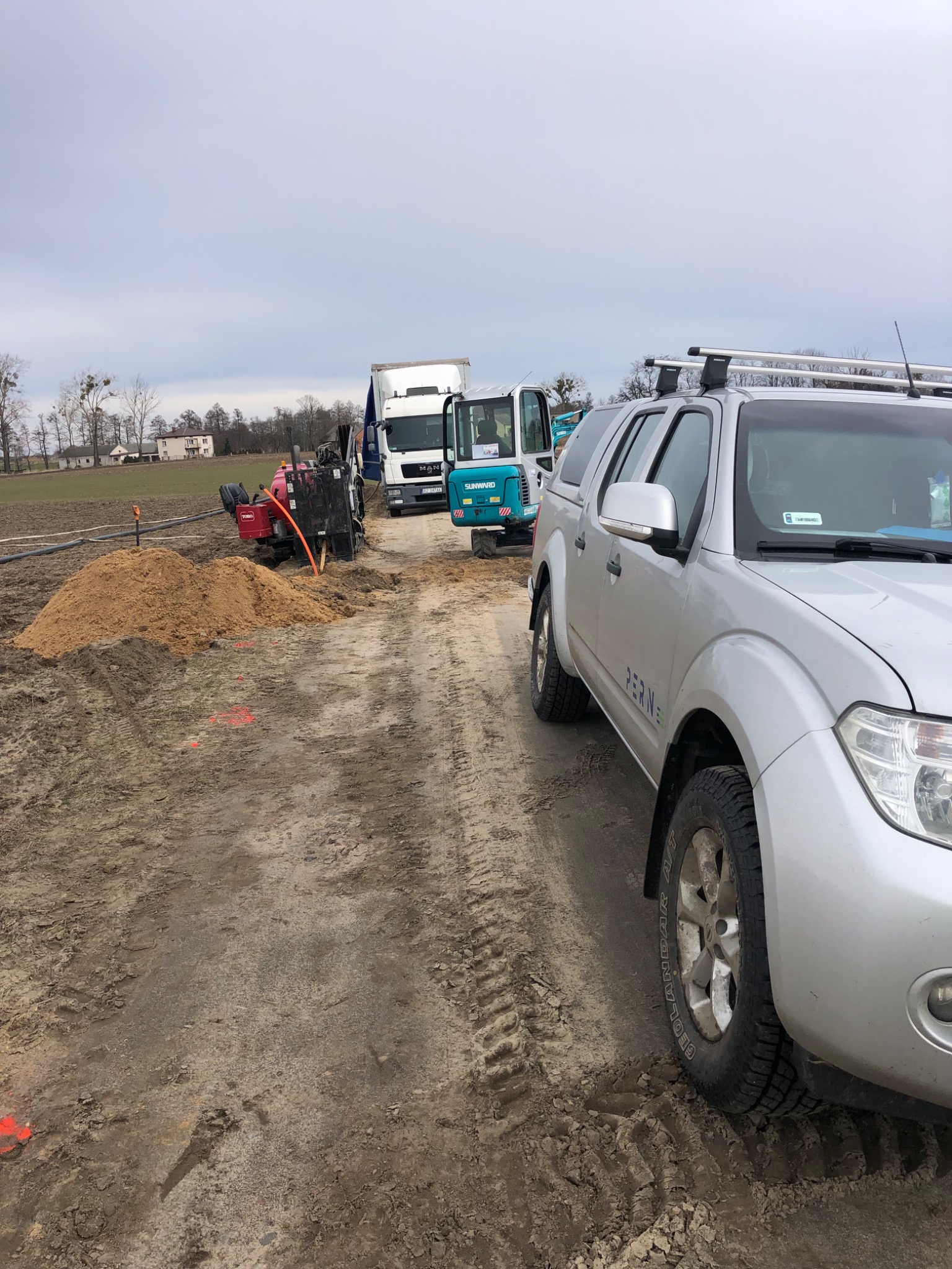 Srebrny samochód terenowy Nissan na placu budowy z mini koparką, ciężarówką MAN i wiertnicą Toro w tle, teren oznaczony pomarańczowymi znacznikami.