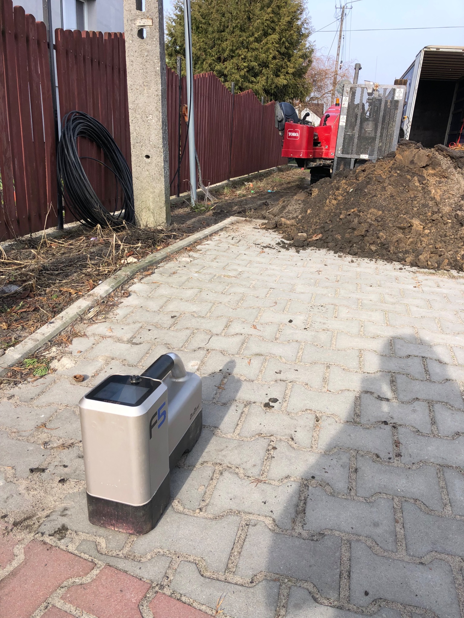 Czerwona mini-koparka Toro na ziemi obok przyczepy, zwój kabli przy drewnianym płocie i wykrywacz kabli F5 na kostce brukowej.