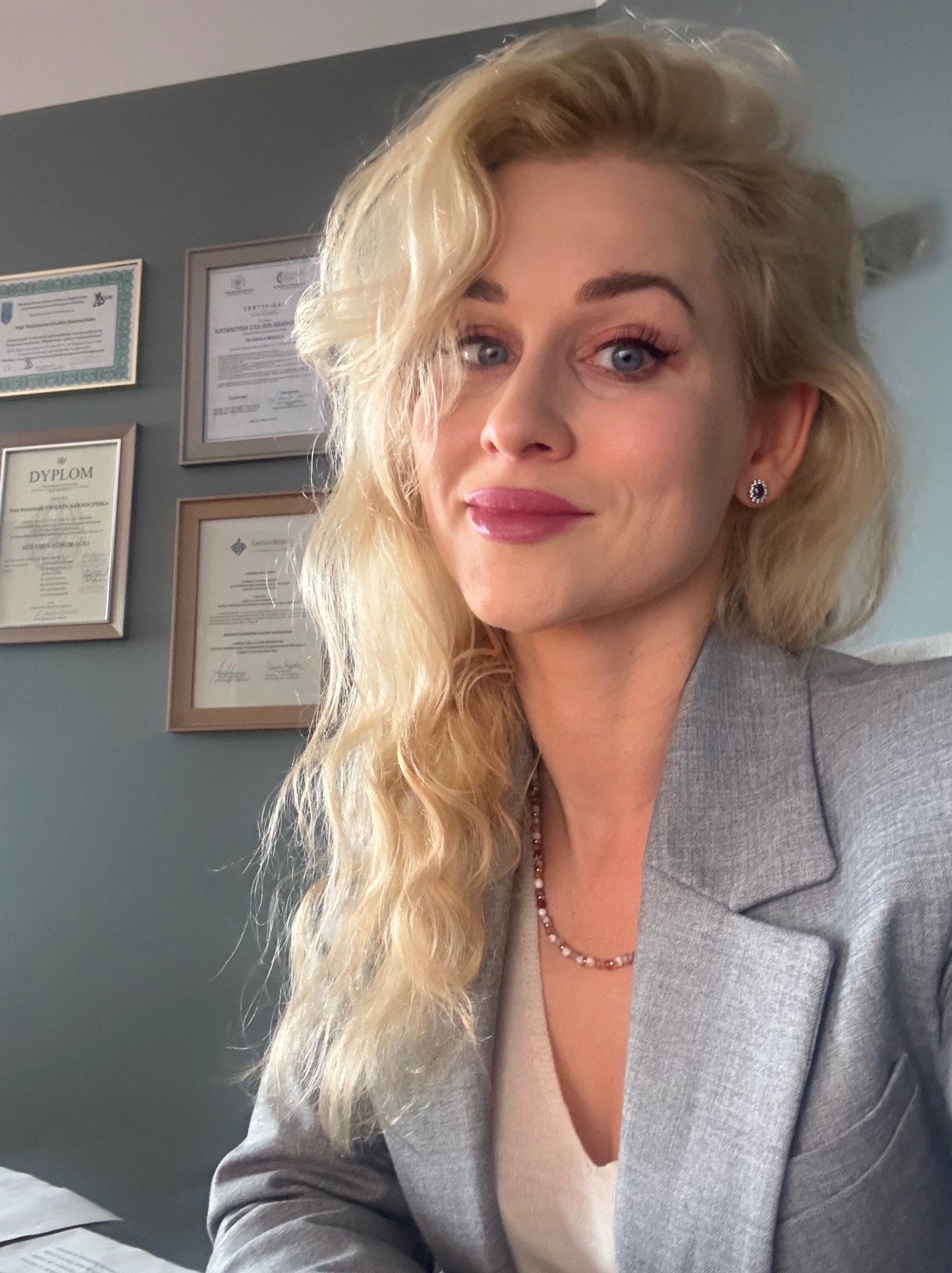 Portret uśmiechniętej kobiety w szarym żakiecie, z blond włosami i dyplomami w tle, sugerujący profesjonalizm w branży prawnej.