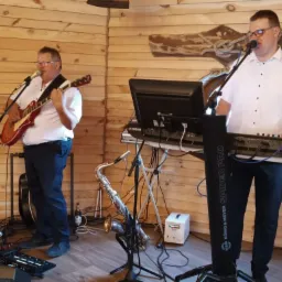 Dwóch mężczyzn grających muzykę na żywo: jeden na gitarze elektrycznej, drugi na keyboardzie, z saksofonem stojącym obok keyboardu, na tle drewnianej ściany.