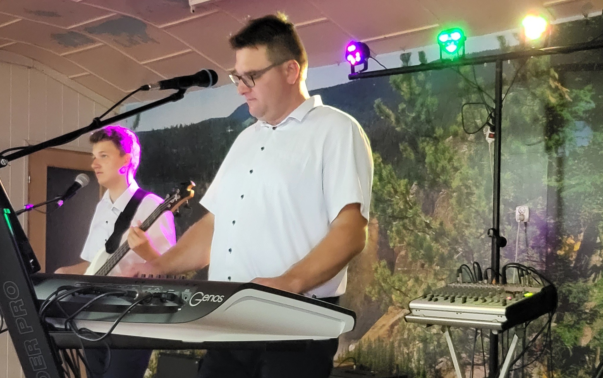 Dwaj muzycy w białych koszulach grający na keyboardzie Genos i gitarze basowej na tle ściany z motywem leśnym, oświetleni kolorowymi reflektorami.