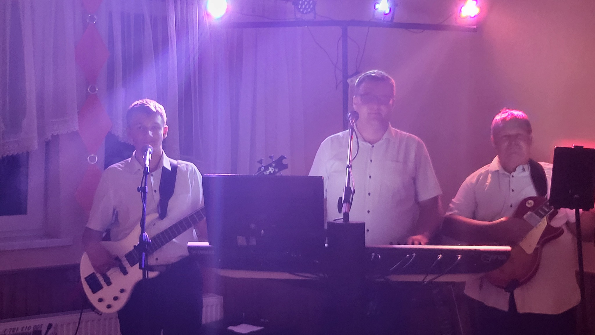 Trzech muzyków grających na instrumentach podczas występu na scenie oświetlonej fioletowymi światłami; jeden z gitarą basową, drugi gra na keyboardzie, a trzeci na gitarze elektrycznej.