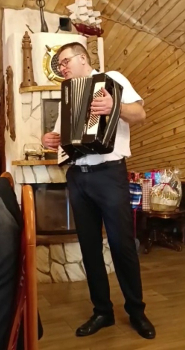 Mężczyzna w okularach gra na akordeonie w rustykalnym wnętrzu z drewnianą boazerią i kominkiem z dekoracjami marynistycznymi.