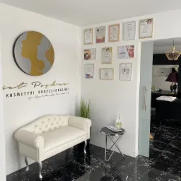 Recepcja salonu kosmetycznego z białym pikowanym siedziskiem, stolikiem kawowym, logiem salonu na ścianie, certyfikatami i czarną marmurową podłogą.