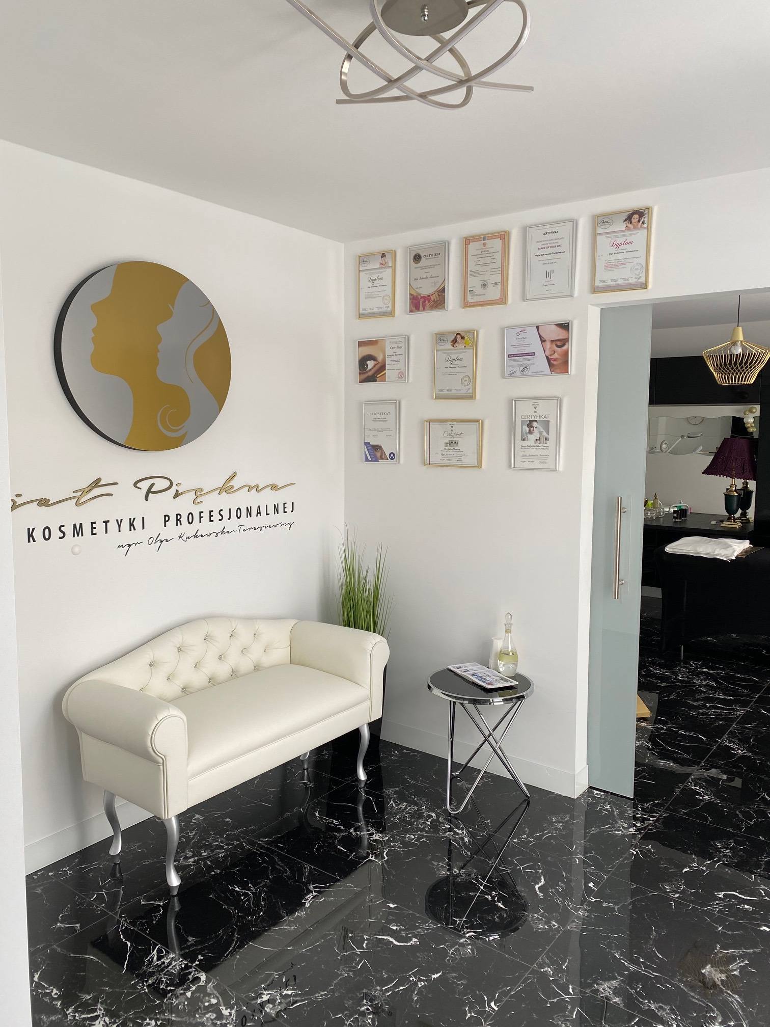 Recepcja salonu kosmetycznego z białym pikowanym siedziskiem, stolikiem kawowym, logiem salonu na ścianie, certyfikatami i czarną marmurową podłogą.