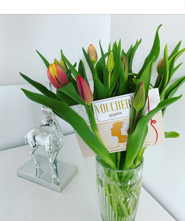 Bukiet czerwono-żółtych tulipanów w szklanym wazonie na białym stoliku, obok srebrna figurka jelenia, voucher salonu kosmetycznego w tle.