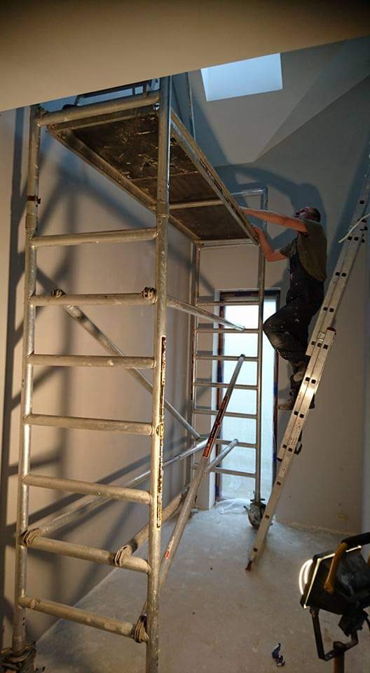 Pracownik na rusztowaniu i drabinie w trakcie prac wykończeniowych, widoczne surowe ściany, okno dachowe i lampa budowlana na podłodze.