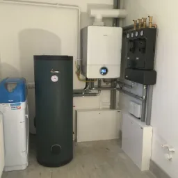 Zestaw urządzeń grzewczych i uzdatniania wody w kotłowni: biały piec gazowy Bosch z wyświetlaczem, zasobnik Kospel, stacja uzdatniania wody Akwa 3 oraz armatura na ścianie, całość połączona rurami.