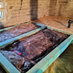 Konstrukcja drewniana z izolacją z wełny mineralnej, częściowo przykryta płytą OSB, w tle ceglana ściana z oknem, wiertarka akumulatorowa i miara zwijana na płycie, piła ręczna z czerwonym uchwytem...