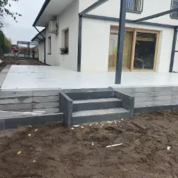 Wykończony taras z jasnych płyt, z trzema stopniami wejściowymi z szarej kostki, otoczony ziemią i widocznym fragmentem domu z pergolą.